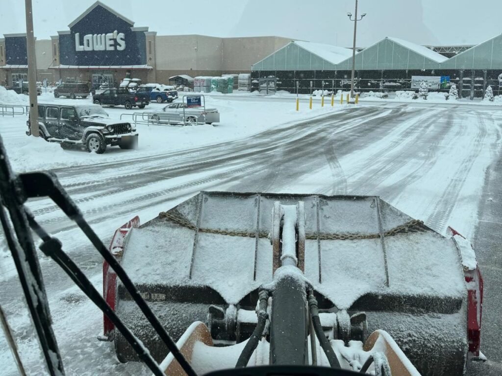 View from a wheel loader cab clearing a big-box retail parking lot during active snowfall